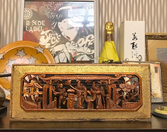 May include: A wooden carved panel with a gold background and a scene of people in a garden. The panel is framed in a gold frame. The panel is on a wooden shelf with other items.