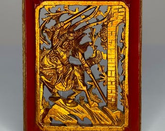May include: A rectangular, gold-colored, carved relief artwork depicting a warrior on horseback, set within a red frame. The intricate design features a detailed battle scene with a brick wall and foliage.