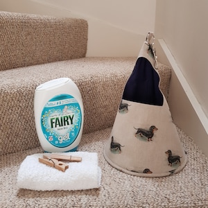 May include: A blue and white bottle of Fairy Non Bio Gel washing up liquid, a white towel with two wooden clothes pegs, and a beige fabric storage basket with a blue lining and a dachshund print.