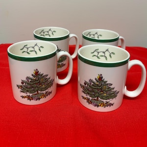 May include: Four white ceramic mugs with a Christmas tree design. Each mug features a green rim and handle, with a detailed illustration of a decorated Christmas tree on the front. The mugs are set on a red surface.