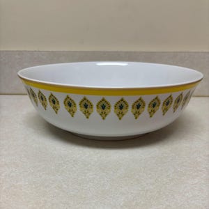 May include: White ceramic serving bowl with a yellow and gold rim. The bowl features a repeating pattern of yellow and navy blue floral designs around the exterior. The bowl is sitting on a beige surface.