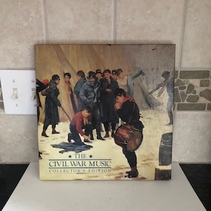 May include: A collector's edition of "The Civil War Music" album cover. The cover art depicts a group of soldiers in a winter setting, with a drummer in the foreground. The album cover is rectangular and has a vintage aesthetic.