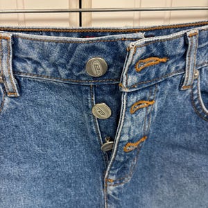 May include: Close-up of a pair of blue denim jeans. The jeans feature a button-fly closure with silver buttons and orange stitching details. The denim has a faded wash, and the image is well-lit.
