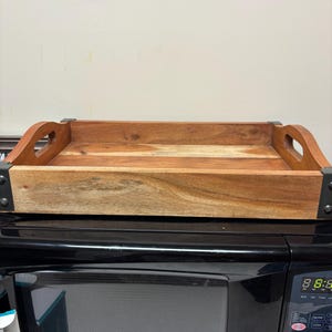 May include: A rectangular wooden serving tray with cut-out handles. The tray is made of light brown wood with darker wood grain and metal corner accents. The tray is sitting on top of a black microwave.
