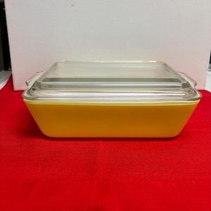 May include: A rectangular yellow Pyrex baking dish with a clear glass lid. The dish has rounded corners and handles on each side. The lid sits flush on top of the dish. The dish is on a red surface.