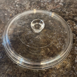 May include: A clear glass Pyrex lid with a small, rounded handle. The lid is circular and has a slightly raised rim. The handle is also clear and rounded. The lid is designed for cookware.