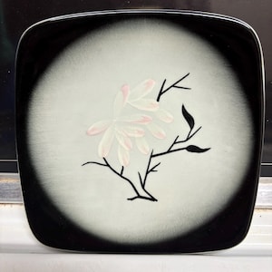 May include: Square black and white decorative plate with a floral design. The plate features a black border, a white center, and a pink and black floral illustration. The plate is likely used for serving food or as a decorative item.