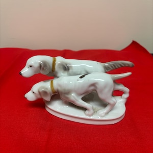 May include: A white porcelain figurine featuring two hunting dogs with gray markings and yellow collars. The dogs are posed in a dynamic stance, mounted on a white oval base. The figurine is set against a red background.