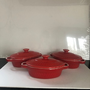 May include: Three red ceramic casserole dishes with lids. Each dish has a round shape with a ribbed design and small handles. The lids have a matching ribbed design and a small knob. The dishes are on a white surface.