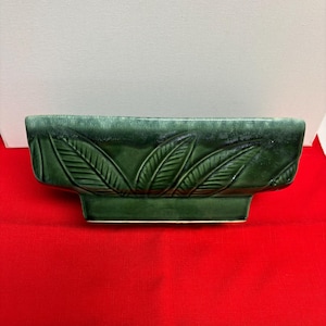 May include: A rectangular, dark green ceramic planter with a raised base. The planter features a repeating leaf design along the front. The glossy finish and the dark green color give it a vintage look. The planter is suitable for indoor or outdoor use.