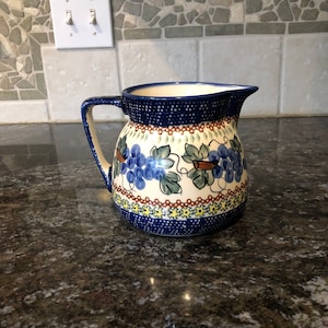 May include: A blue and white ceramic pitcher with a grapevine design. The pitcher has a brown handle and a wide mouth.