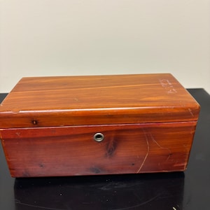 May include: A rectangular wooden box with a reddish-brown finish. The box has a hinged lid and a small, round keyhole. The wood grain is visible, and the box appears to be well-crafted. The box is likely used for storage.