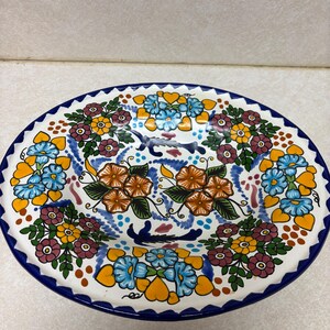 May include: A white ceramic plate with a blue rim and intricate floral designs in orange, yellow, blue, and maroon. The plate features a scalloped edge and is likely handmade, showcasing a vibrant, artistic style.