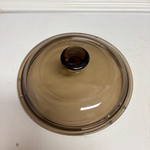 May include: A round, amber-colored glass lid with a dark brown knob. The lid is designed for cookware and has a transparent appearance, allowing visibility of the contents. The lid is made of heat-resistant glass.