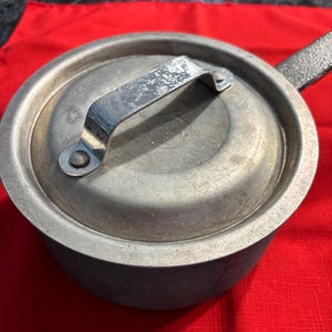 May include: A small, vintage, silver-colored saucepan with a lid and handle. The lid has a curved handle and a circular design in the center. The saucepan is sitting on a red surface.
