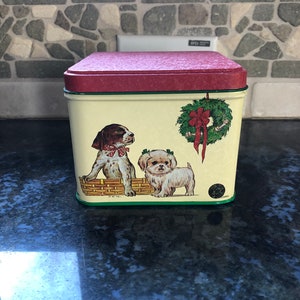 May include: A rectangular metal tin with a maroon lid and green trim. The tin features illustrations of two puppies, one in a basket and the other standing, along with a Christmas wreath. The background is a cream color.