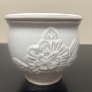 May include: A white ceramic pot with a rounded body and a flared rim. The exterior features a raised, embossed floral design with detailed petals and leaves. The surface has a glossy finish, and the base is slightly tapered toward the bottom.