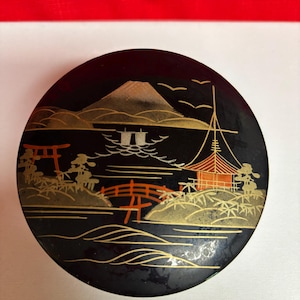 May include: A round, black decorative box with a Japanese landscape design. The design features a mountain, bridge, and buildings in gold and red against a black background. The box has a glossy finish.