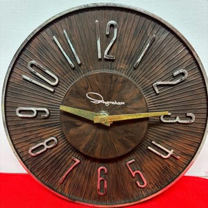 May include: A vintage round clock with a brown and silver face. The clock features silver numerals and hands, with the brand name "Ingraham" in cursive. The clock's face has a radial design, and the hands are gold.