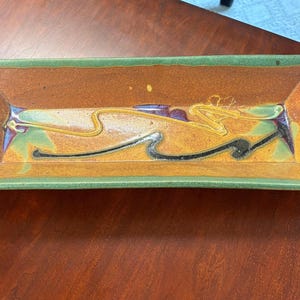 May include: Rectangular ceramic serving tray with a brown interior and green rim. The tray features abstract yellow, black, and purple designs. The tray has handles on each end.