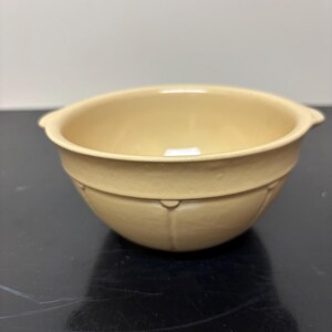 May include: A light beige ceramic bowl with a smooth, matte finish. The bowl features a rounded shape with two small, integrated side handles and a subtle, embossed vertical panel design encircling the lower exterior section of the vessel.