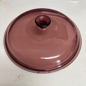 May include: A round, transparent, purple glass lid with a circular design and a small, rounded handle. The lid is designed for cookware and has a slightly raised center with a series of radiating lines.