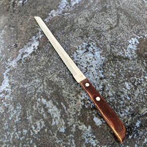 May include: A serrated bread knife with a stainless steel blade and a dark brown wooden handle. The handle is secured with two silver rivets. The knife is designed for slicing bread and other baked goods.