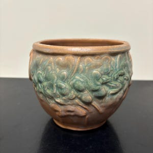 May include: A small, round terracotta pot featuring a textured, raised relief pattern around its center. The relief design is finished in a muted green glaze that contrasts with the natural, earthy brown tone of the unglazed clay body.