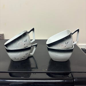 May include: Four white and black teacups and saucers are stacked on a black surface. The cups have a black rim and handle, with a floral design. The saucers are white with a black rim. The cups and saucers are arranged in two stacks.