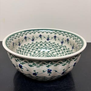 May include: A round ceramic bowl with a scalloped exterior edge, featuring a cream-colored base decorated with a repeating green lattice pattern and small blue floral accents arranged in a delicate vine motif around the interior and exterior.