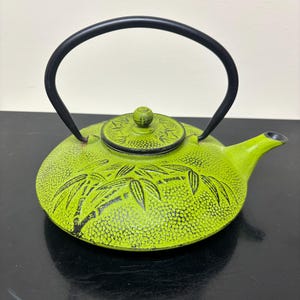 May include: A bright lime green cast iron teapot featuring a textured hobnail surface and a raised black bamboo leaf design. The teapot includes a matching lid with a small knob and a curved black metal handle attached to the sides.