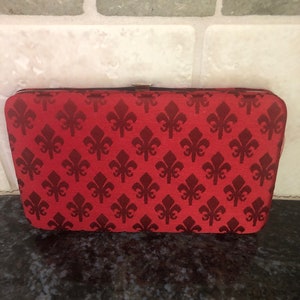 May include: A rectangular red clutch with a repeating dark red fleur-de-lis pattern. The clutch has a metal clasp and a black trim. The background is a light-colored brick wall.