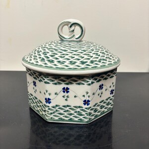 May include: A hexagonal ceramic lidded jar featuring a white base with a green lattice pattern on the lid and base. The center of the jar displays a delicate hand-painted vine motif with small blue four-petaled flowers and green leaves.
