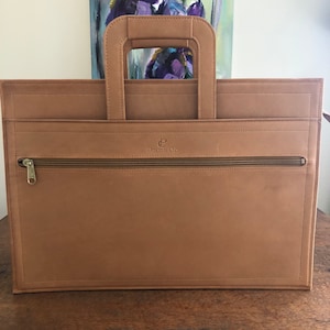 May include: A tan leather briefcase with a top handle and a zippered front pocket. The briefcase has a rectangular shape and a stitched border. The brand name "Penhaligon's Club" is visible on the front.