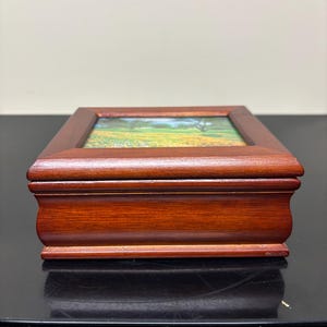 May include: A rectangular wooden jewelry box with a hinged lid. The box is stained a rich, dark brown. The lid features a framed landscape painting of a field of yellow flowers, green grass, and trees. The box sits on a black surface.