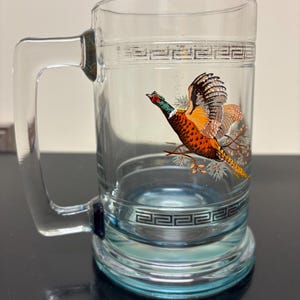 May include: Clear glass beer mug with a handle and a decorative pheasant design. The mug features a blue base and a black and white geometric pattern around the top and bottom. The pheasant is depicted in flight with vibrant colors.