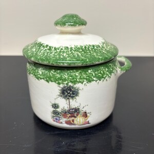 May include: A white ceramic lidded jar featuring a green sponge-painted rim and lid. The body displays a colorful illustration of a potted topiary, a bird's nest, and a yellow striped teapot. The jar includes a small side handle for easy gripping.