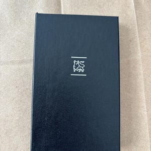May include: A rectangular, black box with a textured surface. A small, white emblem of a bull is centered on the front, above two horizontal lines. The box is closed and appears to be made of a sturdy material.