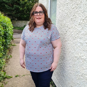 May include: A woman wearing a white and blue striped shirt with red bows printed on it. She is wearing blue jeans and glasses.