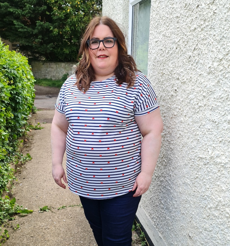 May include: A woman wearing a white and blue striped shirt with red bows printed on it. She is wearing blue jeans and glasses.