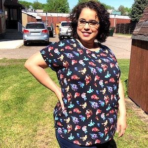 May include: A woman wearing a black short-sleeved shirt with a colorful floral print. The shirt has a round neckline and a relaxed fit. She is wearing blue jeans and has her hair pulled back.