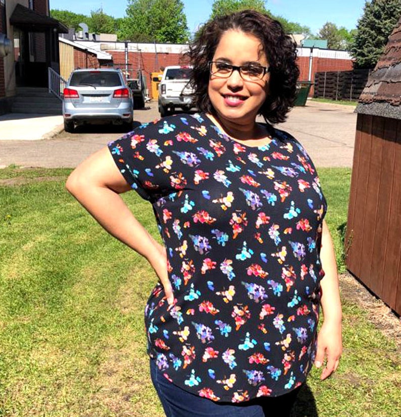 May include: A woman wearing a black short-sleeved shirt with a colorful floral print. The shirt has a round neckline and a relaxed fit. She is wearing blue jeans and has her hair pulled back.