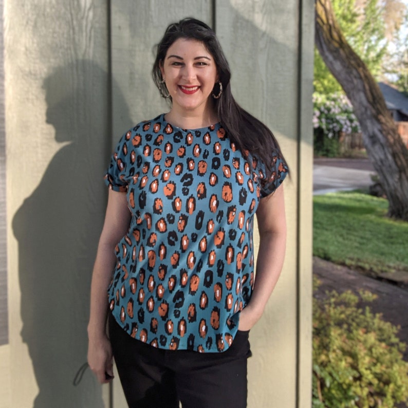 May include: A woman wearing a teal blue and orange leopard print top with short sleeves. She is standing in front of a light green wall.