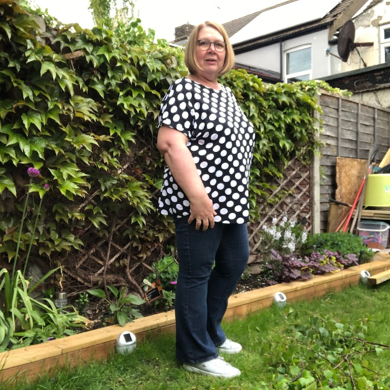 May include: A woman wearing a black and white polka dot shirt with short sleeves and blue jeans. She is standing in a garden with green grass and a wooden fence.