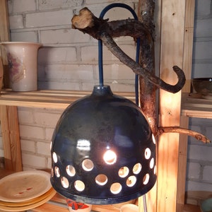 Op de afbeelding: Een blauwe keramische hanglamp met een cirkelvormig ontwerp en gaten erdoorheen. De lamp hangt aan een tak met een blauwe kabel.