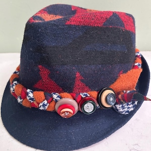 May include: A fedora-style hat with a dark blue brim and crown. The crown features a red and orange geometric pattern. A braided band with buttons and a plaid accent encircles the hat.