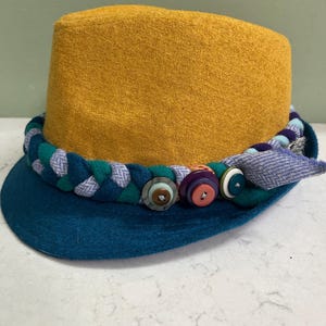 May include: A mustard yellow hat with a teal brim and a decorative braided band. The band is teal, purple, and white, and is adorned with colorful buttons. The hat is a unique fashion accessory.