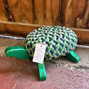 May include: A handmade turtle-shaped footrest. The turtle's body is a mix of green, purple, and gray braided fabric. The base and legs are painted green. The footrest is 25 inches long and 11 inches wide.