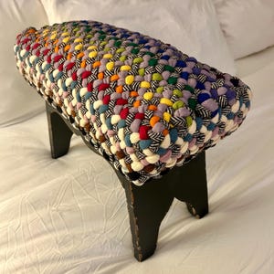 May include: A handcrafted, rectangular footstool with a woven top in a rainbow of colors. The stool's legs are black, and the woven top features a mix of vibrant hues. This decorative piece adds a pop of color to any room.