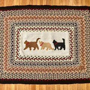 May include: Rectangular braided rug with a cat theme. The rug features a central light blue rectangle with three cat silhouettes in brown, tan, and black. The rug has a brown, red, and cream braided pattern.